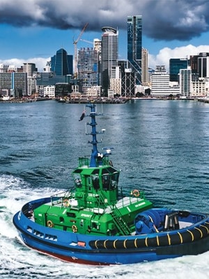 A brightly colored green and blue tugboat advancing on water with a city skyline in the background under a cloudy sky.
